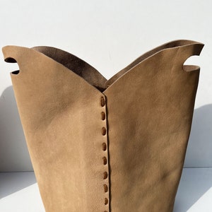 May include: A tan leather storage basket with a stitched seam down the center. The basket has two cut-out handles on the top.