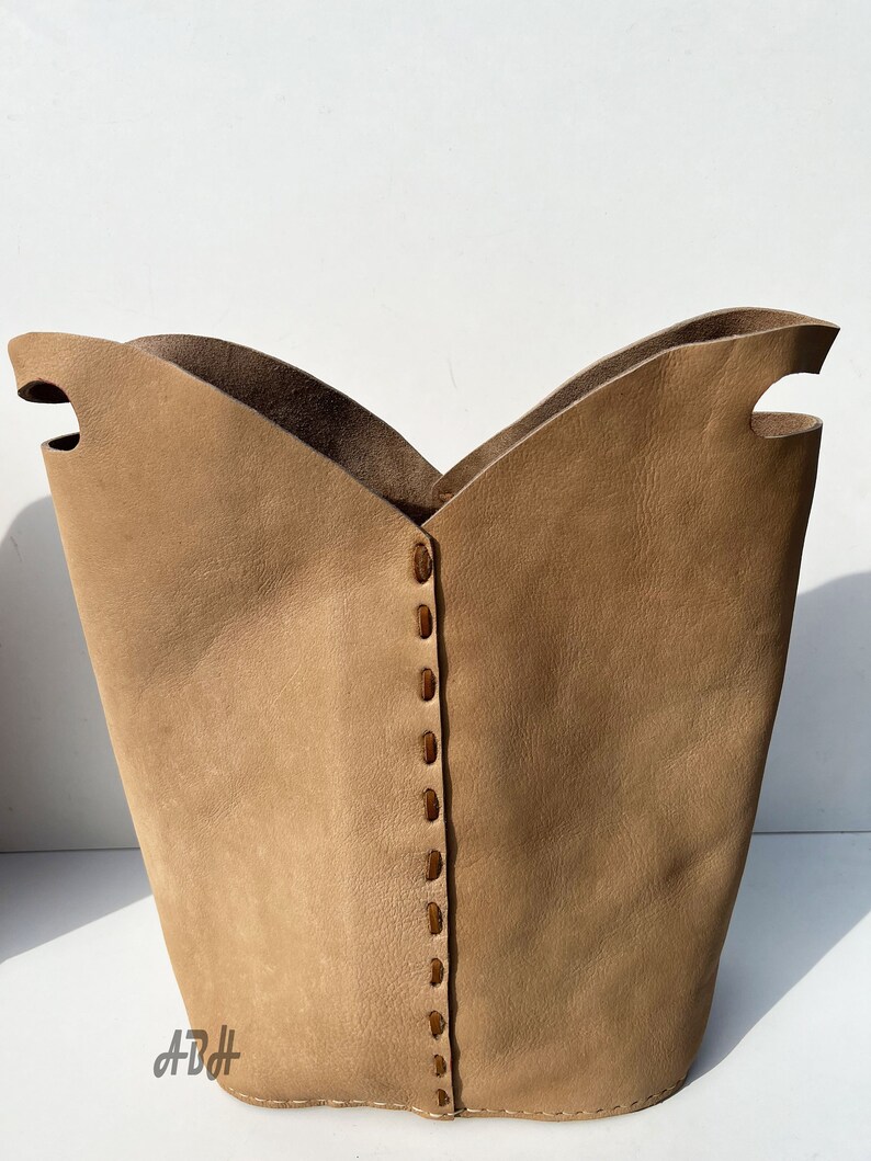 May include: A tan leather storage basket with a stitched seam down the center. The basket has two cut-out handles on the top.