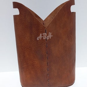 May include: A brown leather storage basket with a stitched seam down the center. The basket has two cut-out handles on the top.
