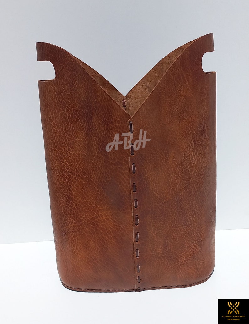 May include: A brown leather storage basket with a stitched seam down the center. The basket has two cut-out handles on the top.