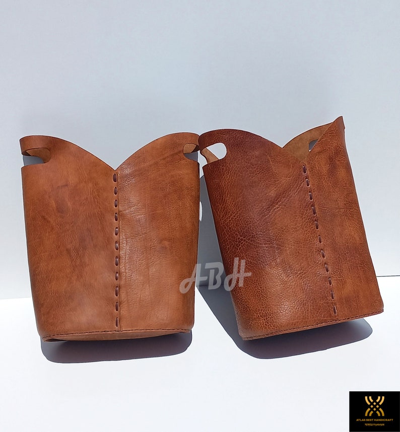 May include: Two brown leather storage baskets with stitched detailing. The baskets are cylindrical in shape and have a wide opening at the top.