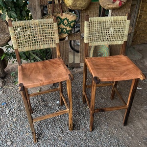 May include: Two wooden bar stools with woven cane backs and brown leather seats. The stools have a rustic, natural look.