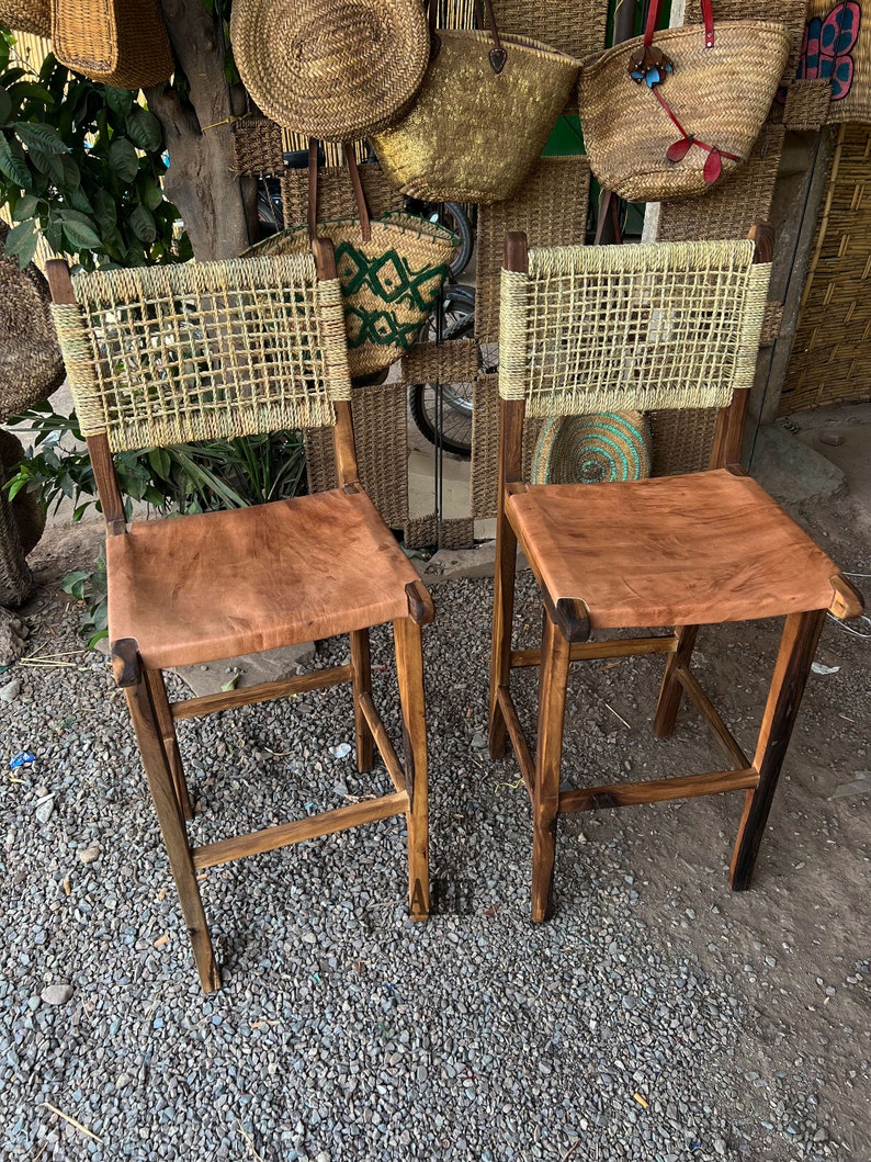 May include: Two wooden bar stools with woven cane backs and brown leather seats. The stools have a rustic, natural look.