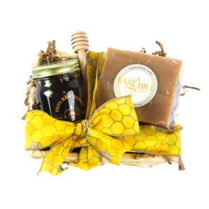 May include: A wicker basket filled with a jar of honey, a bar of soap, and a wooden honey dipper. The soap is labeled "East Hill Honey Co.". The basket is decorated with a yellow bow with a honeycomb pattern.