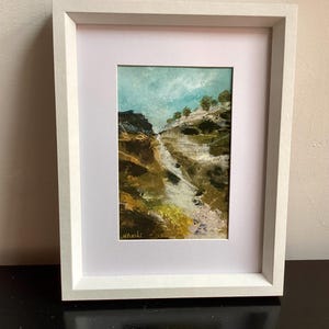May include: An original landscape painting in a white frame. The artwork depicts a mountain scene with a stream, using shades of brown, green, and white. The sky is a light blue. The painting is signed in the lower left corner.