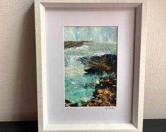 Op de afbeelding: Een ingelijst schilderij van een kustlandschap met turkoois water, donkere rotsachtige oevers en gestructureerde penseelstreken in tinten blauw, wit en oranje. Het kunstwerk is rechtsonder gesigneerd met LH Fields en zit in een witte lijst.