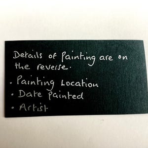 Op de afbeelding: Een rechthoekige zwarte kaart met witte handgeschreven tekst: Details of Painting are on the reverse. Painting Location, Date painted, Artist. De kaart heeft een matte textuur en geeft informatie over het kunstwerk.