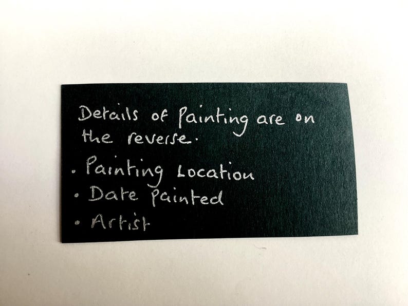 Op de afbeelding: Een rechthoekige zwarte kaart met witte handgeschreven tekst: Details of Painting are on the reverse. Painting Location, Date painted, Artist. De kaart heeft een matte textuur en geeft informatie over het kunstwerk.