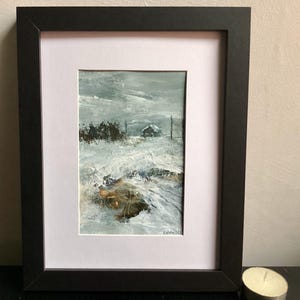 Op de afbeelding: Een olieverfschilderij in een zwarte lijst met een winterlandschap, een klein huisje in de verte, besneeuwde grond en een donkere, bewolkte lucht. Het kunstwerk toont dikke, gestructureerde penseelstreken in tinten wit, grijs en bruin.
