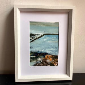 May include: A framed painting of a coastal scene featuring a calm blue pool of water, dark rocky shorelines with orange accents, and a distant horizon line under a grey sky. The artist signature L. Fields is visible in the bottom left corner.