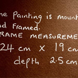 Op de afbeelding: Een donkerbruin rechthoekig oppervlak met witte handgeschreven tekst die luidt: The Painting is mounted and Framed. FRAME MEASUREMENTS: 24 cm x 19 cm depth 2.5 cm. De tekst geeft de fysieke afmetingen van het ingelijste kunstwerk in centimeters weer.
