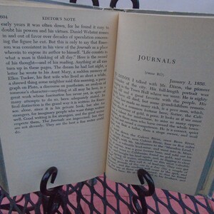 Pu&ograve; includere: Un libro aperto con pagine ingiallite, che mostra una sezione intitolata "Journals (from RO)". Il testo parla di un uomo di nome Mr. Dixon, un pioniere della citt&agrave;, e la sua storia di vita. Il libro &egrave; su un tavolo di metallo con una superficie rosa.
