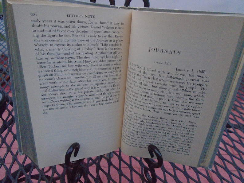 Pu&ograve; includere: Un libro aperto con pagine ingiallite, che mostra una sezione intitolata "Journals (from RO)". Il testo parla di un uomo di nome Mr. Dixon, un pioniere della citt&agrave;, e la sua storia di vita. Il libro &egrave; su un tavolo di metallo con una superficie rosa.