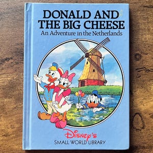 Puede incluir: Un libro de tapa dura azul claro titulado Donald and the Big Cheese: An Adventure in the Netherlands de la Disney's Small World Library. La portada presenta una ilustración circular del Pato Donald, Daisy y un molino de viento.