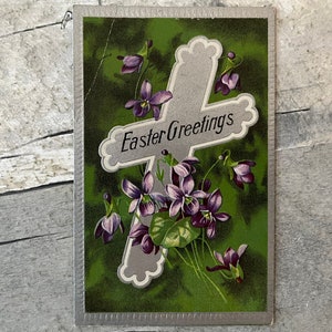May include: A vintage Easter greeting card with a silver cross and purple flowers on a green background. The card reads "Easter Greetings".