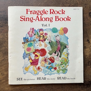 May include: A Fraggle Rock Sing-Along Book Vol. I with a white cover featuring a colorful illustration of five Fraggle characters, balloons, and a banner. The text reads Fraggle Rock Sing-Along Book Vol. I and SEE the pictures HEAR the story READ the book.