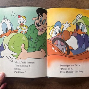 May include: An open children's book featuring colorful illustrations of Donald Duck, Huey, and a man. The left page shows a man handing a green helmet to Donald Duck. The right page shows Donald Duck sitting in a red car wearing the green helmet.