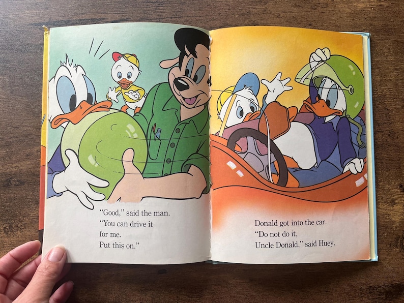 May include: An open children's book featuring colorful illustrations of Donald Duck, Huey, and a man. The left page shows a man handing a green helmet to Donald Duck. The right page shows Donald Duck sitting in a red car wearing the green helmet.