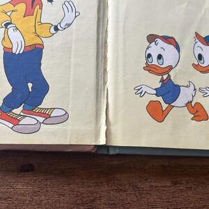 May include: A vintage book page illustration featuring a tall character in a yellow shirt, blue pants, and red sneakers on the left, and two small duck characters wearing blue shirts and orange caps walking on the right against a cream background.