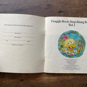May include: An open Fraggle Rock Sing-Along Book Vol. I. The left page features a 'This Fraggle Rock Book Belongs To' section with lines for name, address, city, state, and phone number. The right page shows a colorful illustration of a yellow creature.