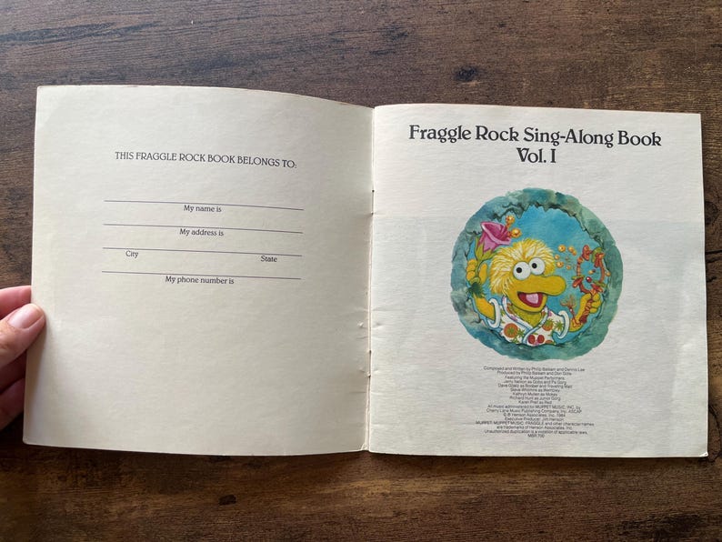 May include: An open Fraggle Rock Sing-Along Book Vol. I. The left page features a 'This Fraggle Rock Book Belongs To' section with lines for name, address, city, state, and phone number. The right page shows a colorful illustration of a yellow creature.