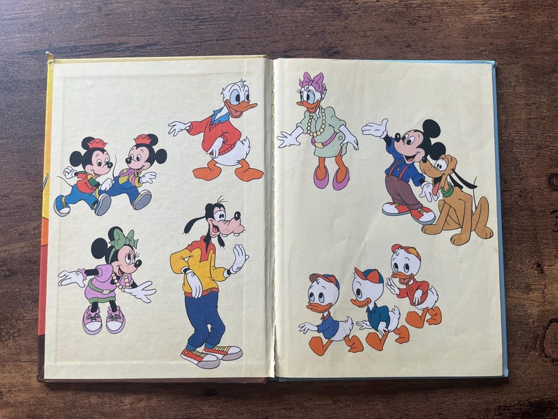 May include: An open book featuring colorful illustrations of classic cartoon characters including Mickey Mouse, Minnie Mouse, Donald Duck, Daisy Duck, Goofy, Pluto, and three ducklings. The characters are depicted in various poses on a light yellow background.