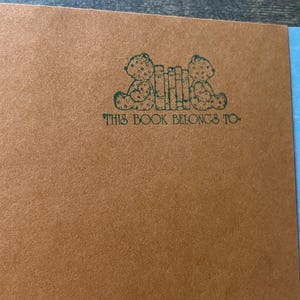 May include: A tan-colored book page featuring a dark green ink stamp of two teddy bears sitting on either side of a row of books. Below the illustration, the text reads, THIS BOOK BELONGS TO, in a dark green, decorative, serif typeface.