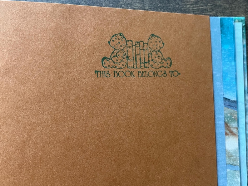 May include: A tan-colored book page featuring a dark green ink stamp of two teddy bears sitting on either side of a row of books. Below the illustration, the text reads, THIS BOOK BELONGS TO, in a dark green, decorative, serif typeface.