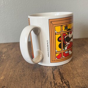 Puede incluir: Taza de cer&aacute;mica blanca con una ilustraci&oacute;n de un personaje de dibujos animados en una ventana. La taza tiene un asa curva y el texto "The Walt Disney Company" impreso en el lateral. La taza est&aacute; sobre una superficie de madera.