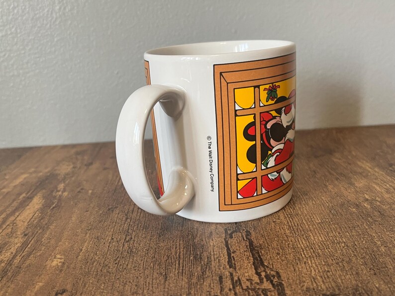 Puede incluir: Taza de cer&aacute;mica blanca con una ilustraci&oacute;n de un personaje de dibujos animados en una ventana. La taza tiene un asa curva y el texto "The Walt Disney Company" impreso en el lateral. La taza est&aacute; sobre una superficie de madera.