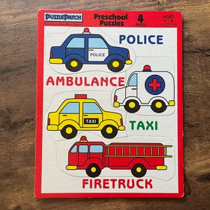 May include: A PuzzlePatch preschool puzzle board featuring four removable vehicle pieces: a blue police car, a white ambulance with a red cross, a yellow taxi, and a red fire truck. The board includes the text labels POLICE, AMBULANCE, TAXI, and FIRETRUCK.