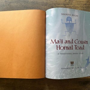 May include: An open hardcover book titled Ma'ii and Cousin Horned Toad by Shonto Begay. The left page is solid orange, while the right page features a light blue background with a dark blue figure, the title text, and Scholastic Inc. branding.