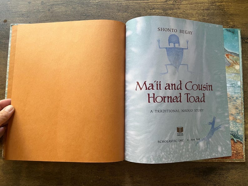 May include: An open hardcover book titled Ma'ii and Cousin Horned Toad by Shonto Begay. The left page is solid orange, while the right page features a light blue background with a dark blue figure, the title text, and Scholastic Inc. branding.