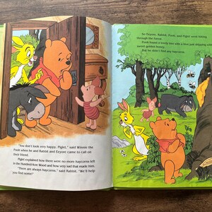 Puede incluir: Un libro de cuentos infantil abierto con Winnie the Pooh, Piglet, Eeyore y Rabbit. La p&aacute;gina izquierda muestra a los personajes en una puerta, mientras que la derecha los representa en un bosque junto a un &aacute;rbol de miel. El texto narra la b&uacute;squeda.
