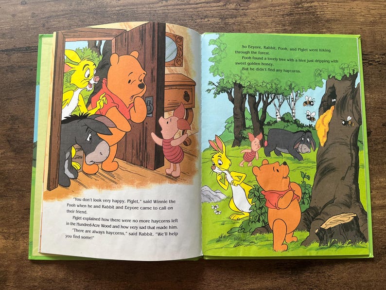Puede incluir: Un libro de cuentos infantil abierto con Winnie the Pooh, Piglet, Eeyore y Rabbit. La p&aacute;gina izquierda muestra a los personajes en una puerta, mientras que la derecha los representa en un bosque junto a un &aacute;rbol de miel. El texto narra la b&uacute;squeda.
