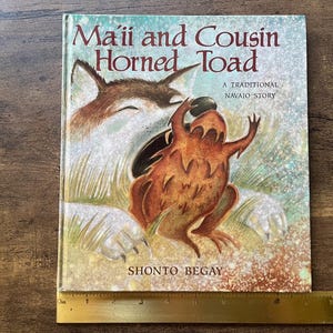 May include: A hardcover book titled Ma'ii and Cousin Horned Toad, A Traditional Navajo Story by Shonto Begay, featuring an illustration of a coyote and a horned toad. A yellow ruler is positioned at the bottom, showing a width of 10 inches.