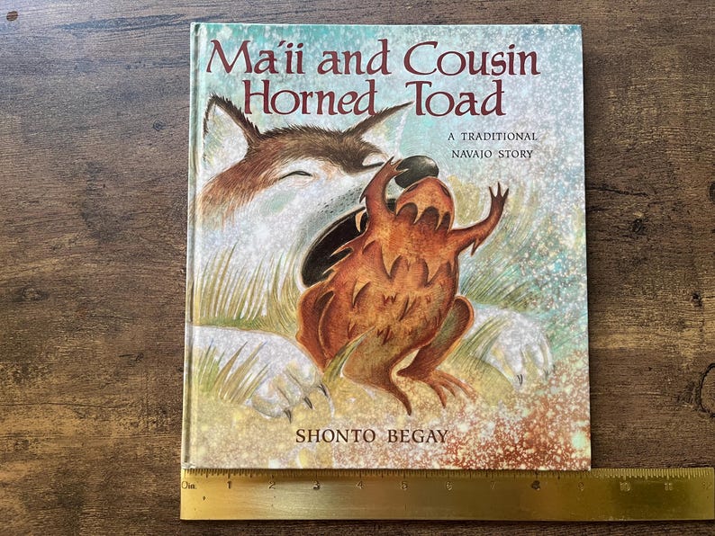 May include: A hardcover book titled Ma'ii and Cousin Horned Toad, A Traditional Navajo Story by Shonto Begay, featuring an illustration of a coyote and a horned toad. A yellow ruler is positioned at the bottom, showing a width of 10 inches.