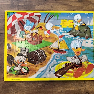 May include: A vintage Disney picture puzzle featuring Donald Duck, Daisy Duck, and nephews Huey, Dewey, and Louie at the beach. The puzzle has a yellow border and depicts a colorful beach scene with a sailboat and a dolphin.
