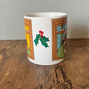 Puede incluir: Taza de cer&aacute;mica blanca con un dise&ntilde;o navide&ntilde;o. La taza presenta ilustraciones de Pap&aacute; Noel, ventanas y acebo. Las ventanas tienen marcos naranjas y el fondo es de color azul claro.