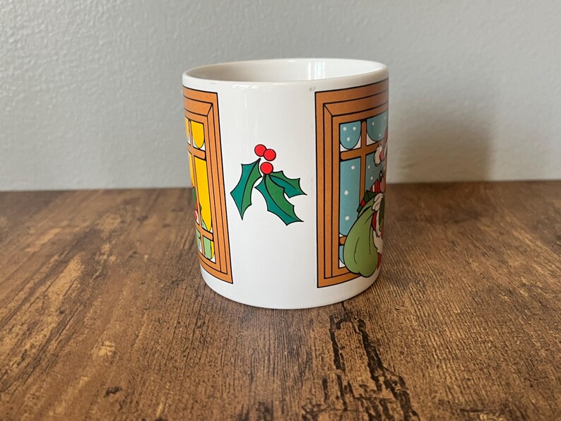 Puede incluir: Taza de cer&aacute;mica blanca con un dise&ntilde;o navide&ntilde;o. La taza presenta ilustraciones de Pap&aacute; Noel, ventanas y acebo. Las ventanas tienen marcos naranjas y el fondo es de color azul claro.