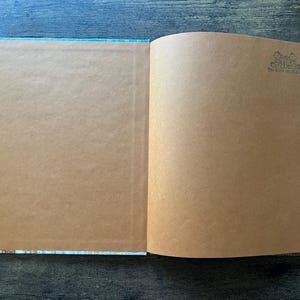 May include: An open book with blank, light brown paper pages. The right page features a small, dark ink stamp in the top right corner that reads THE BOOK THESAURUS. The book has a thin, light blue patterned border visible along the outer edges.