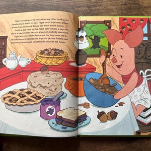 Puede incluir: Una colorida ilustraci&oacute;n de un libro de cuentos con Piglet de Winnie the Pooh sentado a una mesa. Piglet remueve un cuenco azul con una cuchara de madera. La mesa est&aacute; llena de comida, incluyendo un pastel, una tarta, un s&aacute;ndwich y mermelada.