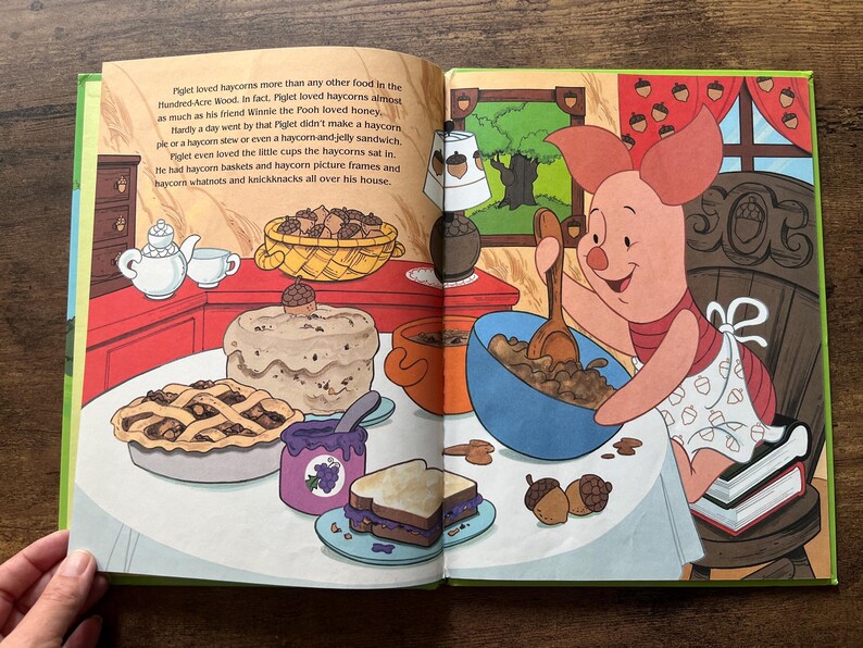 Puede incluir: Una colorida ilustraci&oacute;n de un libro de cuentos con Piglet de Winnie the Pooh sentado a una mesa. Piglet remueve un cuenco azul con una cuchara de madera. La mesa est&aacute; llena de comida, incluyendo un pastel, una tarta, un s&aacute;ndwich y mermelada.
