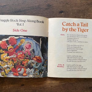 May include: An open Fraggle Rock Sing-Along Book Vol. I. The left page features a colorful illustration of Fraggle characters playing musical instruments. The right page contains the lyrics to the song Catch a Tail by the Tiger, with text.