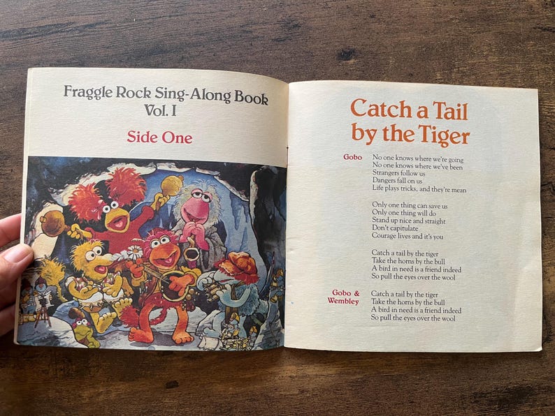 May include: An open Fraggle Rock Sing-Along Book Vol. I. The left page features a colorful illustration of Fraggle characters playing musical instruments. The right page contains the lyrics to the song Catch a Tail by the Tiger, with text.