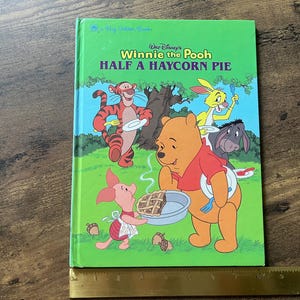 Puede incluir: Un libro de tapa dura verde titulado Walt Disney's Winnie the Pooh Half a Haycorn Pie, con ilustraciones de Pooh, Piglet, Tigger, Rabbit y Eeyore. Una regla de metal est&aacute; colocada en la parte inferior, mostrando una longitud de 25 cm.