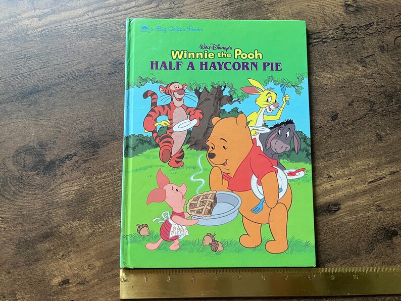 Puede incluir: Un libro de tapa dura verde titulado Walt Disney's Winnie the Pooh Half a Haycorn Pie, con ilustraciones de Pooh, Piglet, Tigger, Rabbit y Eeyore. Una regla de metal est&aacute; colocada en la parte inferior, mostrando una longitud de 25 cm.