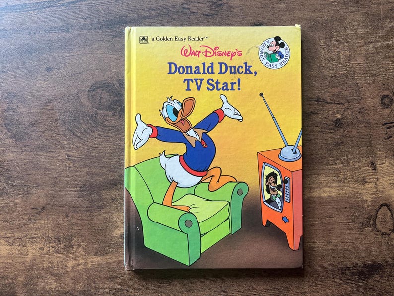 May include: A hardcover children's book titled Walt Disney's Donald Duck, TV Star! featuring a yellow cover with an illustration of Donald Duck standing on a green armchair next to an orange television set with an antenna and a screen showing a character.