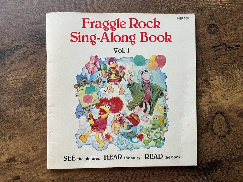 May include: A Fraggle Rock Sing-Along Book Vol. I with a white cover featuring a colorful illustration of five Fraggle characters, balloons, and a banner. The text reads Fraggle Rock Sing-Along Book Vol. I and SEE the pictures HEAR the story READ the book.