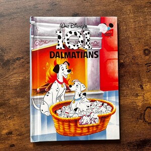 Puede incluir: Un libro de tapa dura titulado Walt Disney's 101 Dalmatians con una colorida ilustración de dálmatas adultos y cachorros en una cesta frente a una chimenea. El título está impreso en letras grandes de color blanco con manchas negras.